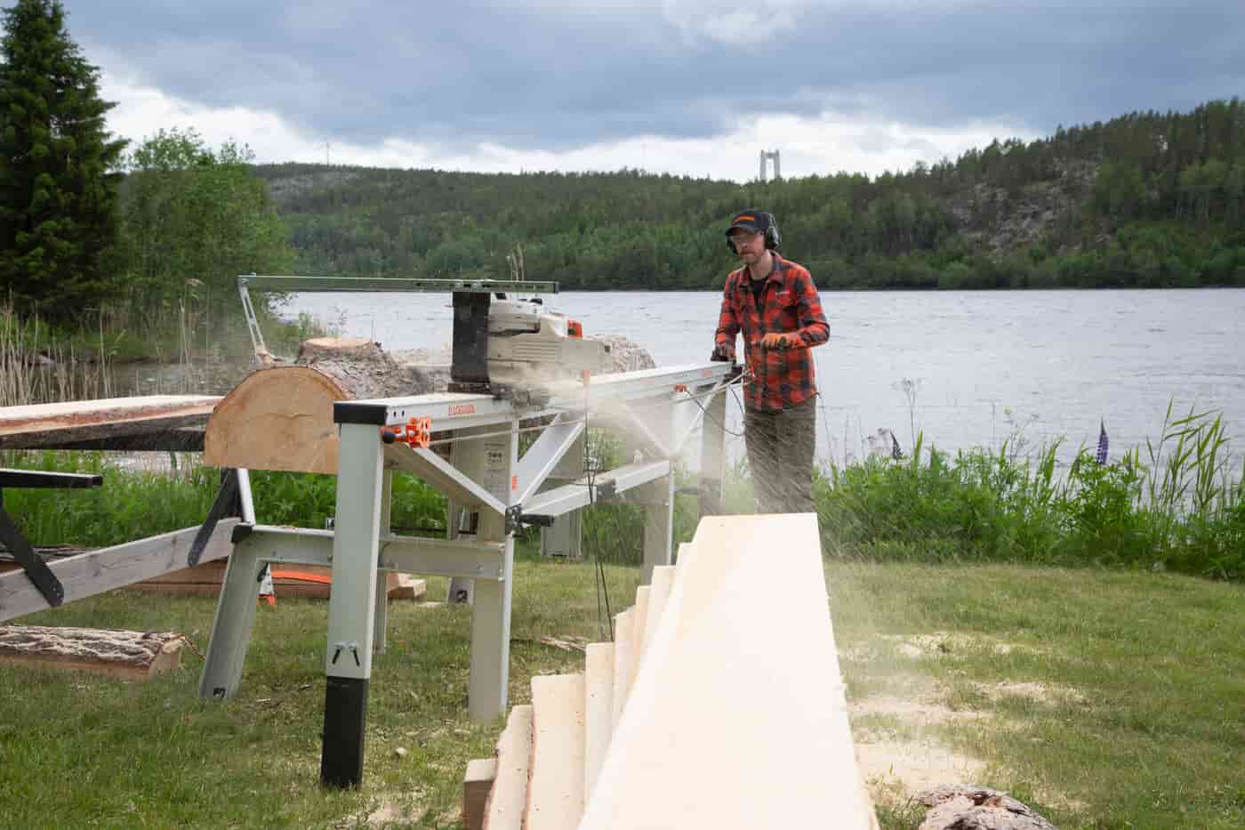 A person sawing a log on a Logosol C701 chainsaw mill beside a lake, with fresh boards in the foreground.